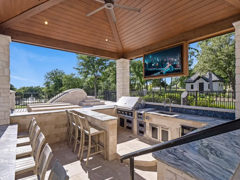 Sunken outdoor kitchen with built-in grill, mounted TV and stone bar seating at Bella Terra Estate, Parker TX