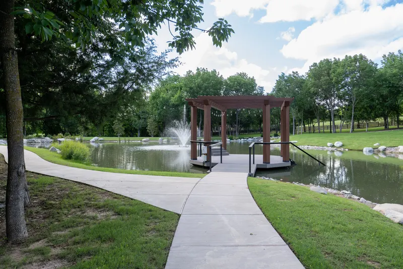 Pond-side dock with cedar pergola and aerating fountain along a paved path at Bella Terra Estate, Parker TX