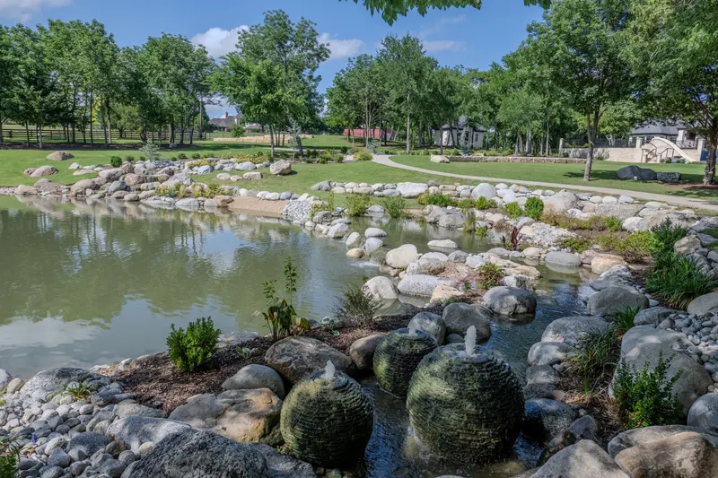 Sphere fountains bubbling among boulders at the pond's edge with the house beyond at Bella Terra Estate, Parker TX