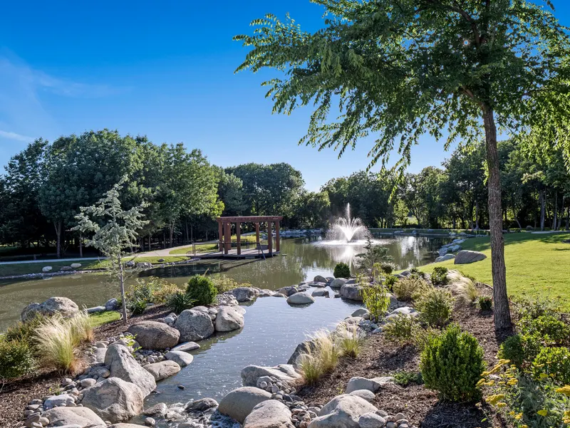 Rock-lined stream spilling toward the pond's tall fountain and cedar dock pergola at Bella Terra Estate, Parker TX