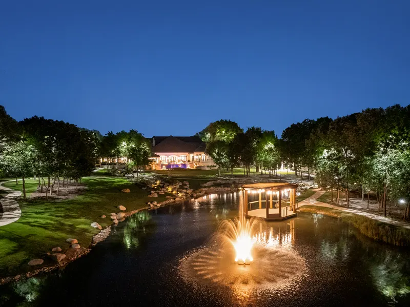 Illuminated pond fountain and dock pergola with the glowing house beyond at Bella Terra Estate, Parker TX