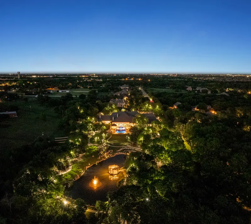 High night aerial of the lit 6.4-acre grounds and pond with distant city lights at Bella Terra Estate, Parker TX