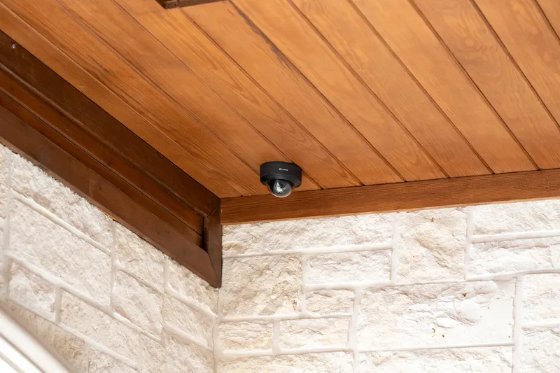Dome security camera tucked under the stained wood soffit above white stone at Bella Terra Estate, Parker TX