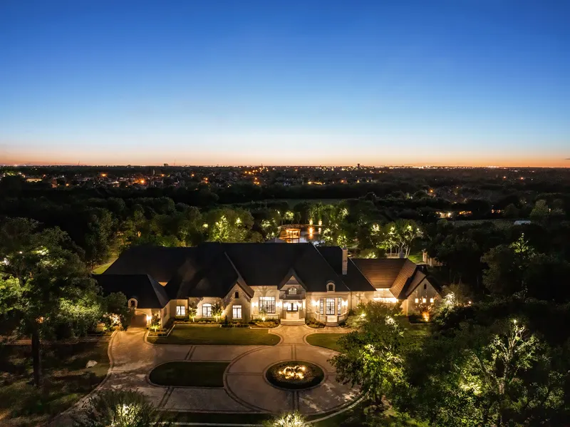 Twilight aerial of the front of Bella Terra Estate with the lit circular drive and Dallas skyline glow, Parker TX